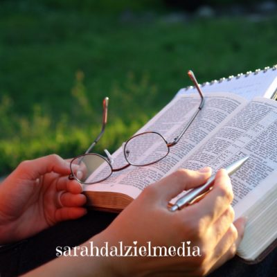 hands of a person holding a pen and glasses over an open bible